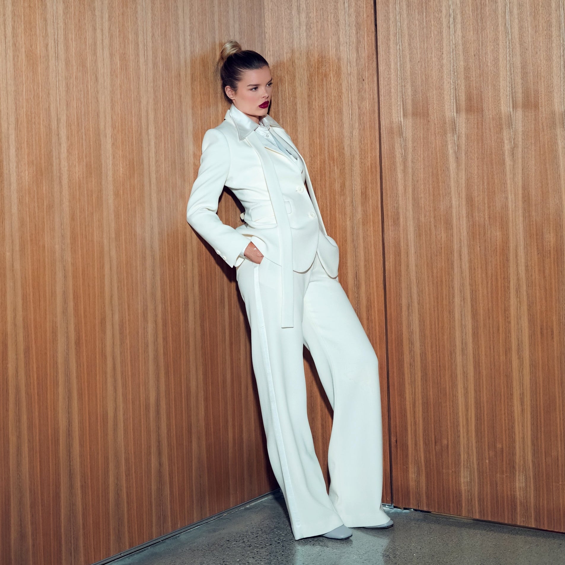 Woman in a white suit standing against a wooden paneled wall.